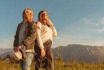 Молодые близкие подруги в стильной одежде стоят вместе на лугу в горах, глядя в золотой свет — стоковое фото