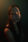 Молодая задумчивая женская модель со светлой проекцией на лице сидит в темной студии и смотрит в сторону — стоковое фото