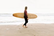 Vista lateral del surfista vestido con traje de neopreno caminando mirando hacia el agua con tabla de surf para coger una ola en la playa durante el amanecer - foto de stock