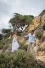 Ponte multirracial encantada e noivo de mãos dadas e andando ao longo da colina arenosa no dia do casamento na natureza — Fotografia de Stock