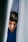 Unglücklicher kleiner Junge, der hinter Vorhängen hervorlugt, während er unter häuslicher Gewalt leidet und sich vor seinen Eltern versteckt — Stockfoto