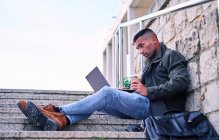 Side view of stylish Hispanic man with coffee to go browsing data on netbook while sitting on steps and working on remote project on city street — Stock Photo