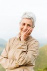 Positive elderly female traveler touching face and looking at camera with smile on blurred background of nature — Stock Photo