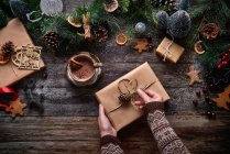 Vue du dessus d'une femme anonyme préparant un cadeau de Noël à côté d'un bol de chocolat et de décorations de Noël sur une table en bois — Photo de stock