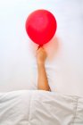 Cropped unrecognizable tired person with red balloon in hand sleeping in bed with white sheets after party — Stock Photo