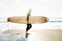 Side view of surfer man dressed in wetsuit walking on the beach with the surfboard in the morning with sunrise in the background — Stock Photo