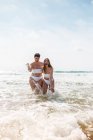 Alegre amigas en trajes de baño abrazándose mientras se paran salpicando agua en el océano espumoso cerca de la playa de arena bajo el cielo azul nublado en el día soleado - foto de stock