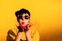 Feminino moderno em hip hop cap e óculos de sol soprando confete colorido e se divertindo contra o fundo amarelo — Fotografia de Stock