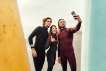 Группа счастливых друзей-серферов, одетых в гидрокостюмы, стоящих рядом с досками для сёрфинга, делая селфи со смартфоном на пляже во время тренировки — стоковое фото