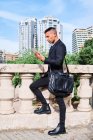 Вид сбоку сфокусированный молодой латиноамериканец исполнительный менеджер в стильном костюме с мешком чтения информации на планшете, стоя рядом перила на городской террасе — стоковое фото