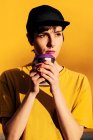 Giovane femmina informale in berretto alla moda sorseggiando take away bevanda calda contro sfondo giallo — Foto stock