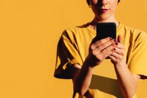 Mujer contemporánea con corte de pelo elegante y piercing usando smartphone en las redes sociales contra el fondo amarillo - foto de stock