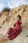 Спокойная женщина в длинном летнем платье сидит на песчаном холме и смотрит в камеру — стоковое фото