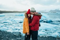 Vue latérale du jeune homme et de la jeune femme en tenue d'hiver avec des caméras professionnelles debout sur la rive près de l'eau avec de la glace — Photo de stock