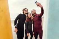 Grupo de amigos surfistas felizes vestidos com fatos de mergulho perto de pranchas de surf enquanto tomam selfie com smartphone na praia durante o treino — Fotografia de Stock