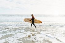 Vista lateral del surfista vestido con traje de neopreno caminando con tabla de surf hacia el agua para coger una ola en la playa durante el amanecer - foto de stock