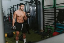 Мускулистый молодой тренер мужского пола с обнаженным туловищем, поднимающим тяжелые гантели во время тренировки в спортзале — стоковое фото