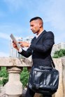 Вид сбоку сфокусированный молодой латиноамериканец исполнительный менеджер в стильном костюме с мешком чтения информации на планшете, стоя рядом перила на городской террасе — стоковое фото