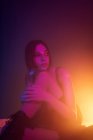 Спокойная молодая модель в платье сидит на полу и опирается на колено, глядя в темную студию с красочными огнями — стоковое фото