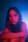 Ruhige junge weibliche Modell in Kleid auf dem Boden sitzend und auf dem Knie gestützt, während Blick in die Kamera in dunklen Studio mit bunten Lichtern — Stockfoto