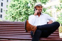 Low angle of smiling self assured young bearded Hispanic gentleman in classy outfit and hat speaking on mobile phone while resting on bench in city — Stock Photo