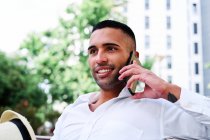 Low angle of smiling self assured young bearded Hispanic gentleman in classy outfit and hat speaking on mobile phone while resting on bench in city — Stock Photo