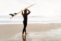Vista lateral do jovem surfista irreconhecível com cabelo comprido vestido de fato de mergulho em pé olhando para a praia com a prancha na cabeça durante o nascer do sol — Fotografia de Stock