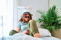 Jeune femme souriante en tenue d'été et chapeau avec des lunettes de soleil assis sur le lit et naviguant sur smartphone tout en passant du temps seule à la maison — Photo de stock