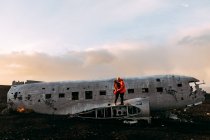 Pareja joven parada en un avión destrozado entre tierras desiertas y el cielo azul - foto de stock