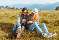 Повне тіло молоді друзі-жінки в капелюхах і стильний одяг, що охолоджується на зеленому газоні і ділиться телефоном в сільській місцевості гір — стокове фото