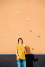 Jovem talentoso macho realizando truque com bolas de malabarismo enquanto em pé no pavimento perto de parede laranja brilhante — Fotografia de Stock