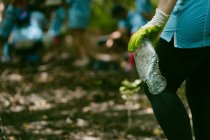 Crop attivista anonimo in guanti protettivi raccogliendo bottiglia di plastica da terra, mentre la raccolta dei rifiuti in natura — Foto stock