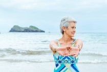 Antiguo pensionista femenina de pelo gris activa en traje de baño elegante estirando los brazos y los hombros mientras hace ejercicios en la playa contra el océano ondulante - foto de stock