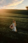 Junge Frau in Vintage-Stil Kleid trägt Weidenkorb beim Spaziergang im grünen Grasfeld bei Sonnenuntergang in der sommerlichen Landschaft — Stockfoto