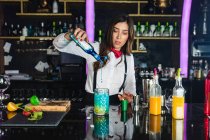 Сфокусований жіночий бармен у стильному вбранні, що додає блакитну рідину з пляшки в склянку, готуючи коктейль, що стоїть за стійкою в сучасному барі — стокове фото
