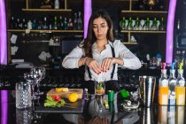 Jeune femme barman en tenue élégante préparant un cocktail mojito avec des tranches de citron tout en se tenant au comptoir dans un bar moderne — Photo de stock