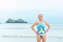 У віці жінка з придатним тілом в стильному барвистому купальнику з геометричним принтом, що стоїть з руками на талії проти моря в літній день — стокове фото