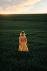 Young female in vintage style looking away thoughtfully while standing alone in grassy field at sunset time in summer evening in countryside — Stock Photo