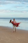 Heureux homme noir portant femme ravie et profitant de l'été sur le bord de mer sablonneux sur fond de mer au coucher du soleil — Photo de stock