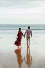 Задний вид многорасовая пара, держась за руки и прогуливаясь вдоль мокрого берега, любуясь морем на закате летом — стоковое фото