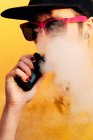 Contemporary female in stylish outfit exhaling fume while standing near yellow wall and vaping on city street — Stock Photo