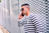 Side view of hispanic man in stylish outfit looking away and talking on cellphone while leaning on wall on city street — Stock Photo