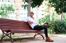 Уверенный в себе молодой латиноамериканец в элегантной одежде и шляпе сидит на скамейке и смотрит в сторону, отдыхая на городской улице — стоковое фото