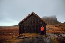 Jovem turista em pé perto da cabana entre terras selvagens perto de altas colinas de pedra e céu nublado — Fotografia de Stock