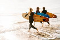 Vista lateral do grupo de amigos surfistas vestidos com fatos de mergulho correndo com pranchas de surf em direção à água para pegar uma onda na praia durante o nascer do sol — Fotografia de Stock
