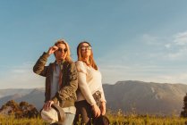 Jeunes amies proches dans des vêtements élégants debout ensemble sur la prairie dans les montagnes regardant loin dans la lumière dorée — Photo de stock