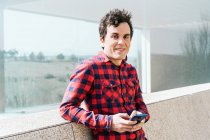 Smiling young male in checkered shirt looking at camera while browsing mobile phone near modern building with glass walls and stone fence — Stock Photo