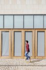 Visão lateral de corpo inteiro do jovem homem ativo em camisa quadriculada e jeans executando truque em monociclo perto de parede de vidro espelhado do edifício contemporâneo — Fotografia de Stock
