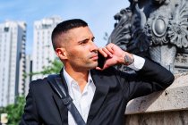 Ganzkörperhispanischer Mann in stylischem Outfit, der wegschaut und mit dem Handy telefoniert, während er sich auf der Stadtstraße an die Wand lehnt — Stockfoto
