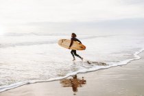 Вид сбоку серфера, одетого в гидрокостюм, бегущего с доской для серфинга к воде, чтобы поймать волну на пляже во время восхода солнца — стоковое фото